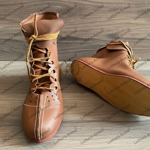 May include: Two brown leather ankle boots with laces. One boot is upright, the other is lying on its side. The boots have a decorative stitch pattern and a red sole. The laces are a lighter brown colour.