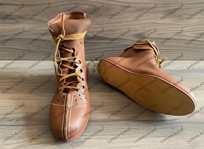 May include: Two brown leather ankle boots with laces. One boot is upright, the other is lying on its side. The boots have a decorative stitch pattern and a red sole. The laces are a lighter brown colour.