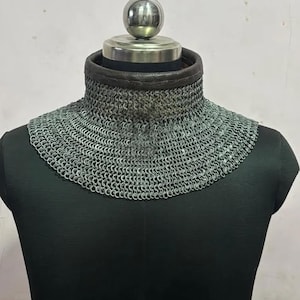 May include: A silver chainmail collar with a dark brown leather trim. The chainmail is made of small, interlocking metal rings. The collar is displayed on a black mannequin.