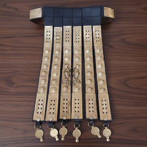 May include: A black leather and brass Roman lorica segmentata apron. The apron features rectangular brass plates with decorative rivets and circular accents, connected by black leather straps. The bottom of the apron has brass pendants.