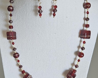 May include: A red beaded necklace and matching earrings set. The necklace features square and oval red beads, interspersed with smaller red and gold beads. The earrings have a pearl accent and dangle with red beads. The jewelry is displayed on a white mannequin.
