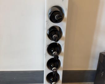 Puede incluir: Un botellero de mármol blanco con un diseño geométrico, que contiene seis botellas de vino tinto. El botellero tiene aberturas circulares para sostener las botellas, mostrando las etiquetas del vino. El mármol tiene vetas grises.