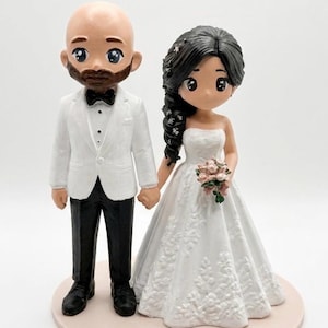 Könnte beinhalten: Ein Tortenaufsatz für Hochzeiten mit zwei nebeneinander stehenden Figuren. Eine Figur ist kahlköpfig mit Bart und trägt ein weißes Sakko, eine schwarze Fliege und eine schwarze Hose. Die andere Figur hat langes dunkles Haar und ein weißes trägerloses Kleid.