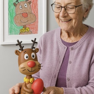 May include: A brown reindeer figurine with a red nose and a yellow collar, holding a red egg. The figurine is on a green base. A framed drawing of a reindeer hangs on the wall behind it.