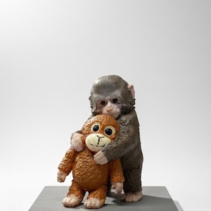 Könnte beinhalten: Eine braune und graue Affenfigur umarmt ein kleineres orangefarbenes Affenplüschtier. Die Figur steht auf einem grauen rechteckigen Sockel vor einem weißen Hintergrund. Das Plüschtier hat ein lächelndes Gesicht.