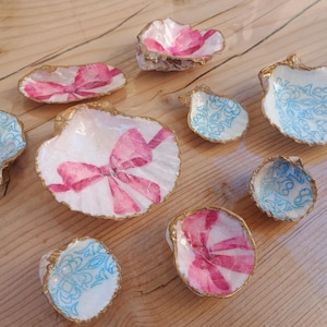 Può includere: Una collezione di conchiglie decorative. Le conchiglie sono dipinte con fiocchi rosa e motivi floreali, e alcune presentano disegni floreali blu. I bordi sono rifiniti con vernice dorata, creando un aspetto delicato ed elegante.