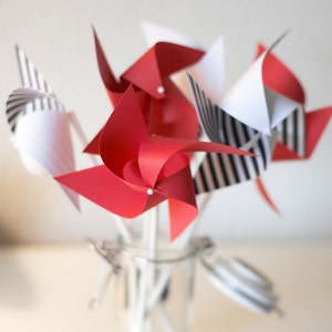 May include: A group of red and black and white paper pinwheels are arranged in a glass jar. The pinwheels are made of paper and have a white stick attached to the back.