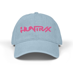 May include: A pale blue baseball cap featuring the pink embroidered text "HUNTRX" across the front. The cap has a curved peak and a classic six-panel construction. The hat is made from a soft, durable material.