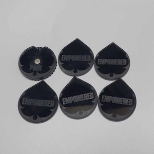May include: A collection of black, teardrop-shaped tokens with the word "EMPOWERED" etched on them. One token has the word "PAIN" and a metal disc. The tokens are arranged on a light surface.