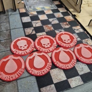 Op de afbeelding: Een set van zeven ronde rode tokens met verhoogde witte afbeeldingen en tekst. Drie tokens tonen een schedelpictogram met het woord CONDEMNED, terwijl vier tokens een druppelpictogram hebben met het woord CONSECRATED in reliëf onderaan.