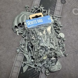 May include: A clear plastic bag labeled BITS BAG containing a collection of unpainted gray plastic miniature model parts, including various limbs, weapons, and armor components, scattered alongside additional loose gray plastic model pieces.