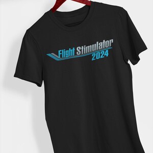 May include: Black t-shirt featuring the text "Flight Simulator 2024" in blue and grey. The design includes a stylized graphic. The shirt is displayed on a wooden hanger.