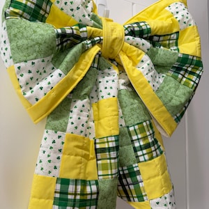 May include: A large, quilted bow decoration in shades of yellow and green, with a patchwork design. The bow features squares of solid yellow, green plaid, and white fabric with green shamrock patterns. The bow is hanging on a white wall.