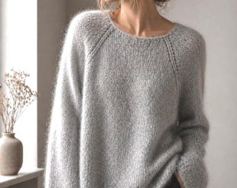 May include: A light gray long-sleeved sweater with a fuzzy, mohair-like texture, a round neckline, and raglan sleeves. The garment features a loose, relaxed fit with a straight hemline and visible knit detailing along the shoulder seams.