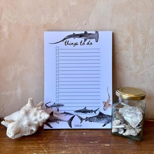 May include: A white rectangular notepad with the text things to do, featuring illustrations of various sharks including a hammerhead and tiger shark, with lined paper and checkboxes for tasks, accompanied by a large seashell and a glass jar of shells.