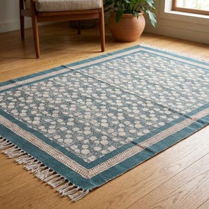 Puede incluir: Una alfombra rectangular con borde turquesa y estampado floral crema. La alfombra tiene un borde con flecos y está sobre un suelo de madera. La alfombra es un elemento decorativo para el hogar. Mide aproximadamente 150 cm x 200 cm.