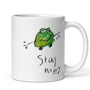 May include: White ceramic mug with a hand-drawn green frog and the words "Stay hoppy" in black script. The frog has a whimsical design with a branch extending from its body. A fun and cheerful design for a coffee or tea mug.