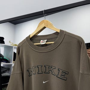 May include: A brown Nike sweatshirt with the brand name in a dark green, arched font across the chest. A small white Nike swoosh logo is below the text. The sweatshirt is hanging on a wooden hanger.