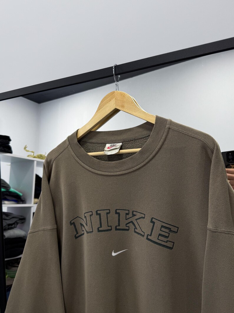May include: A brown Nike sweatshirt with the brand name in a dark green, arched font across the chest. A small white Nike swoosh logo is below the text. The sweatshirt is hanging on a wooden hanger.