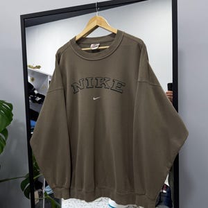 May include: An olive green Nike sweatshirt with the brand name arched across the chest in black lettering. A small Nike swoosh logo is below the brand name. The sweatshirt is hanging on a wooden hanger.