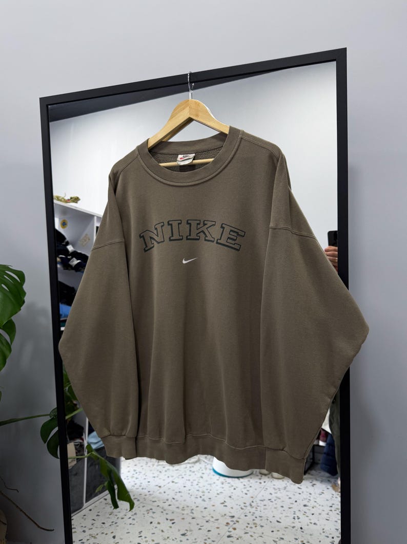 May include: An olive green Nike sweatshirt with the brand name arched across the chest in black lettering. A small Nike swoosh logo is below the brand name. The sweatshirt is hanging on a wooden hanger.