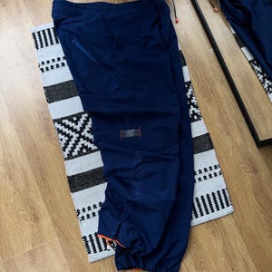May include: Navy blue athletic pants with orange accents. The pants have a drawstring waist and ankle cuffs. A small zipper pocket is visible on the upper left leg. The pants are lying on a black and white patterned rug.