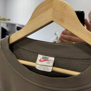 May include: A brown Nike sweatshirt with a crew neck, hanging on a wooden hanger. The tag reads "Nike" in white lettering on a red background, with "Made in USA" below. The sweatshirt is a solid color.