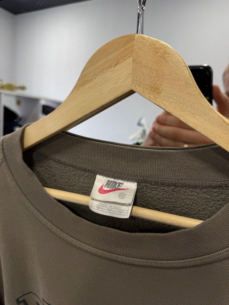 May include: A brown Nike sweatshirt with a crew neck, hanging on a wooden hanger. The tag reads "Nike" in white lettering on a red background, with "Made in USA" below. The sweatshirt is a solid color.