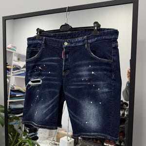 May include: Dark wash denim shorts with distressed details, including a ripped knee and paint splatter accents. The shorts are hanging on a black hanger in front of a mirror, showcasing a casual style.