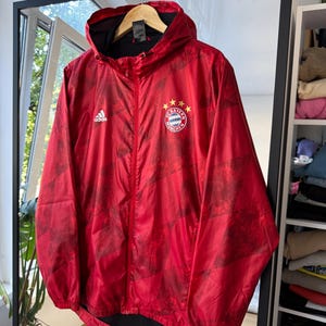Könnte beinhalten: Rote Adidas Kapuzenjacke mit durchgehendem Reißverschluss und dem Bayern München Teamlogo auf der Brust. Die Jacke hat ein schwarzes Innenfutter und ein geometrisches Muster. Das Adidas-Logo befindet sich auf der linken Brust.