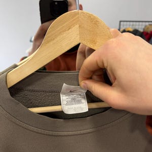 May include: A brown crew neck sweatshirt with the brand name "NIKE" in a dark green color. The sweatshirt is hanging on a wooden hanger. A tag with the Nike logo and text is visible. The tag says "MADE IN USA".