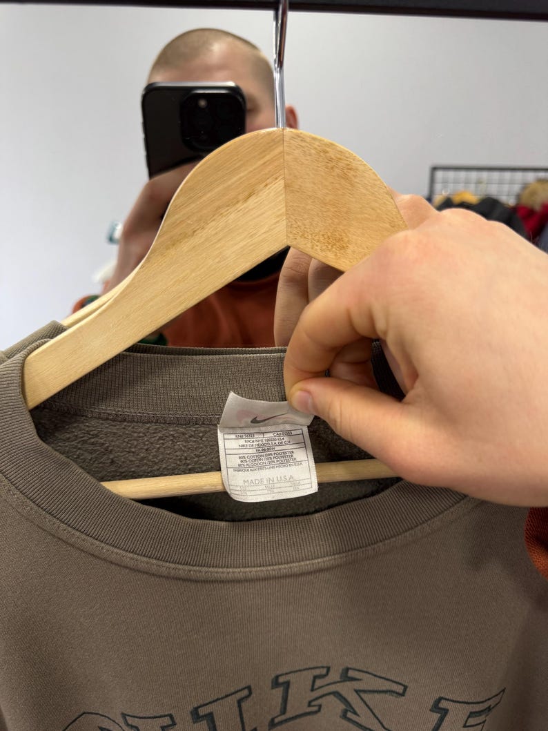 May include: A brown crew neck sweatshirt with the brand name "NIKE" in a dark green color. The sweatshirt is hanging on a wooden hanger. A tag with the Nike logo and text is visible. The tag says "MADE IN USA".