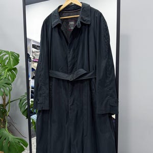 Puede incluir: Un abrigo largo negro con cinturón, colgado de una percha de madera. El abrigo tiene cuello y puños abotonados. El abrigo se exhibe en un marco de metal negro con un espejo.