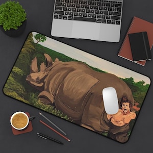 May include: A rectangular mouse pad featuring a painting of a brown rhinoceros lying in a green field. A white computer mouse is on the pad. A man is emerging from the rhino's rear. A laptop, notebook, and coffee cup are also visible.