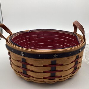Peut inclure: Un panier rond tressé composé de lamelles de bois naturel beige, agrémenté de bandes verticales rouges et bleu marine. Le bord est une bande bleu marine ornée de petites étoiles métalliques argentées et de deux anses en cuir marron.