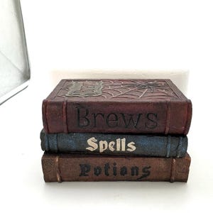 May include: A stack of three rectangular decorative boxes designed to resemble old books. The top box is dark red with a spiderweb and spider relief, labeled 'Brews'. The middle box is blue, labeled 'Spells'. The bottom box is brown, labeled 'Potions'.
