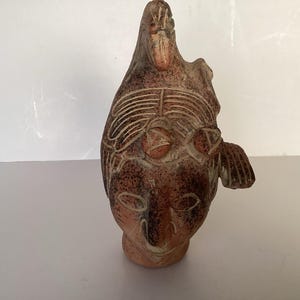 Op de afbeelding: Een terracotta sculptuur met een menselijk hoofd bekroond door een vogelfiguur. Het oppervlak is gevlekt met donkerbruine en roodbruine tinten, met uitgesneden lineaire details die de gelaatstrekken, de vleugels van de vogel en het verenkleed definiëren.