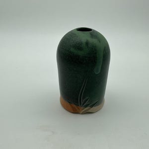 May include: A cylindrical ceramic vase with a rounded top and a small circular opening. The body features a dark forest green glaze with a lighter green drip effect, while the base reveals a natural terracotta clay colour with carved leaf details.