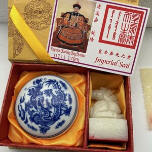 Puede incluir: Una caja de regalo que contiene una tapa de porcelana azul y blanca con un motivo de dragón y un sello de piedra blanca con una figura animal tallada. La tapa muestra una imagen del emperador Qianlong y el texto Imperial Seal y 1711-1799.