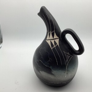 May include: A matte black ceramic pitcher with a rounded body, a curved spout, and a side handle. The surface features etched white line designs, including geometric patterns near the spout and a landscape scene with clouds at the base.