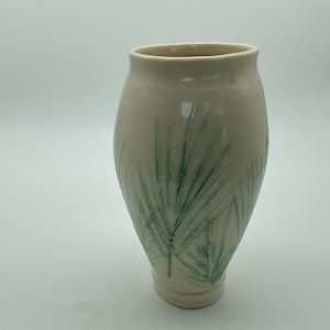 Könnte beinhalten: Eine cremefarbene Keramikvase mit verjüngtem Boden und einer weiten, offenen Oberseite. Die Außenfläche zeigt ein handgemaltes oder geätztes grünes botanisches Muster, das an dünne, längliche Palmwedel oder Grashalme erinnert.