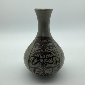 Könnte beinhalten: Eine hellgraue Keramikvase mit engem Hals und bauchigem Körper. Die Oberfläche zeigt ein dunkelbraunes, handgemaltes Grafikdesign, das ein stilisiertes Gesicht mit markanten Augen, einer Nase und einem breiten Mund auf dem runden Boden darstellt.