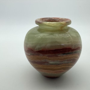 May include: A rounded stone vase featuring horizontal bands of color. The upper section is a pale translucent green, while the lower body displays layered striations of deep red, brown, tan, and white, creating a smooth, polished finish.