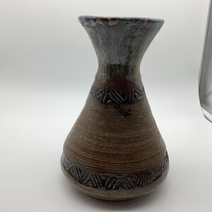 以下が含まれることがあります： 広がった口縁と先細りの首を持つ陶器製の花瓶。本体は水平な溝がある質感のある茶色の仕上げで、濃い青色の幾何学模様が刻まれた帯が2本施されています。首の上部と口縁は濃い青色の釉薬で覆われています。