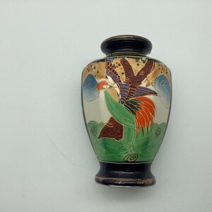 May include: A small ceramic vase with a dark blue neck and base. The body features a hand-painted design of a bird with red, orange, and purple feathers, set against a cream background with green ground, blue clouds, and gold-colored accents.