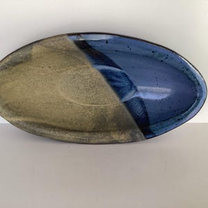 May include: An oval ceramic dish featuring a two-tone glaze. One side has a textured, matte beige finish, while the other side is coated in a glossy, deep blue glaze with subtle dark speckles. The two colours meet in a diagonal line across the centre.