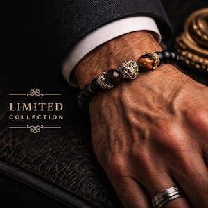 Può includere: Un braccialetto di perline con perline nere opache, un ciondolo centrale a forma di testa di leone color oro e due perline occhio di tigre marroni con accenti dorati. Il testo LIMITED COLLECTION è visibile a sinistra.
