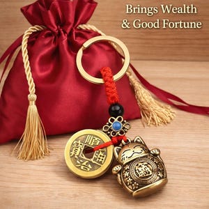 May include: A gold-colored Lucky Cat keychain with a red cord, black bead, and blue accent. The keychain includes a gold coin and a red satin pouch with a gold drawstring. Text reads "Lucky Cat Keychain" and "Brings Wealth & Good Fortune."