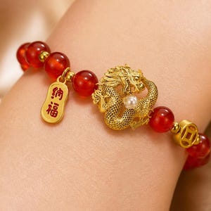May include: A gold-colored bracelet featuring a detailed dragon design with a pearl accent. The bracelet includes red beads and a gold charm with Chinese characters. A gold coin-shaped bead is also present.