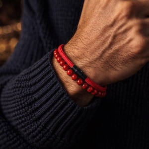 May include: Two red bracelets are worn on a person's wrist. One bracelet features small, round beads, while the other has a woven design. Both bracelets are red and have a black clasp. The person is wearing a dark blue sweater.